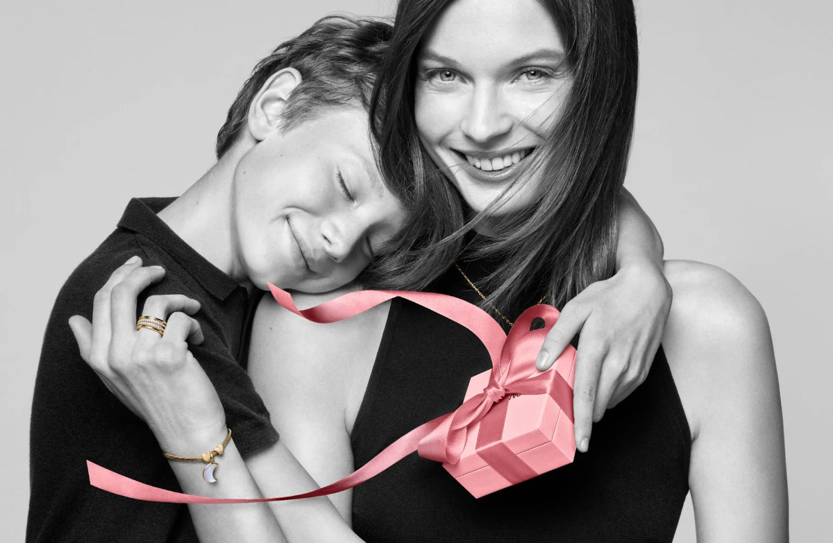 Two women hugging with a pink gift box between them, on a gray background